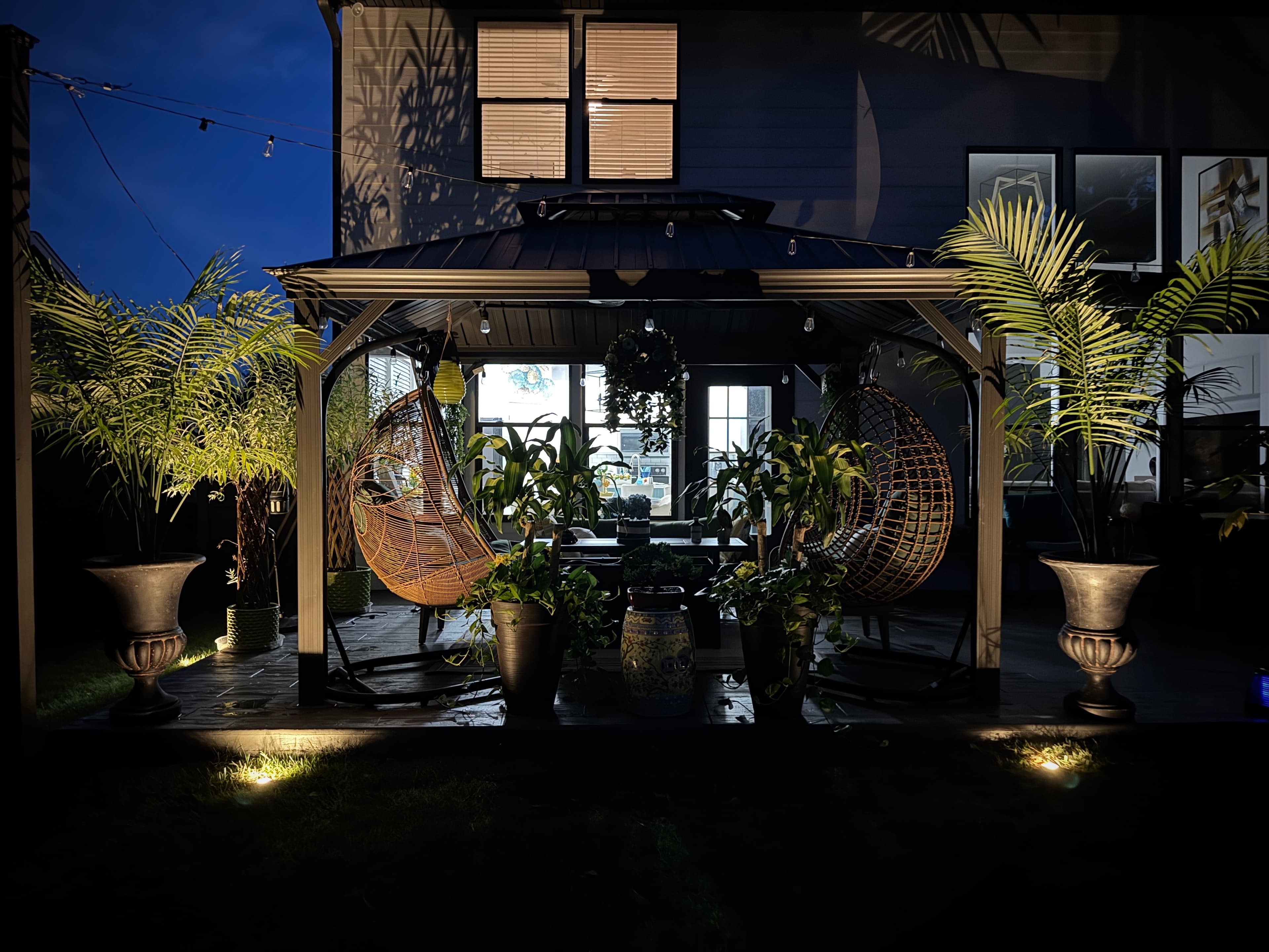 Covered Patio with Hanging Chairs and Ambient Lighting Outdoor patio at twilight with dual hanging wicker chairs, illuminated palm planters, and string lights overhead